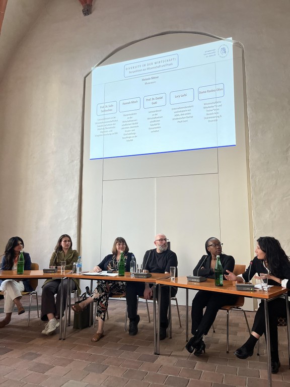 Das Foto zeigt die Panelist*innen der Podiumsdiskussion "Diversity in der Wirtschaft: Perspektiven aus Wissenschaft und Praxis" vom 05.07.23. Von links nach rechts: Prof.in Dr.in Sulin Sardoschau, Hannah Nitsch, Melanie Bittner, Prof. Dr. Daniel Guhl, Lucy Larbi und Dome Ravina Olivo. Über den Panelist*innen ist eine Powerpointfolie an die Wand geworfen mit dem Titel der Diskussion und den Namen (mit Spezialisierungen) in der Sitzreihenfolge. 